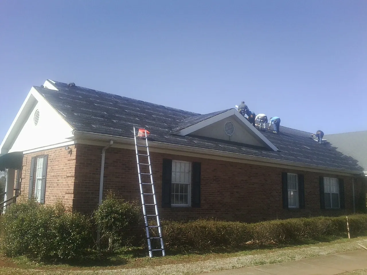 Crew tearing shingles off a brick ranch roof with a ladder set — Emergency Roof Repair work by our crew in Hughes Springs