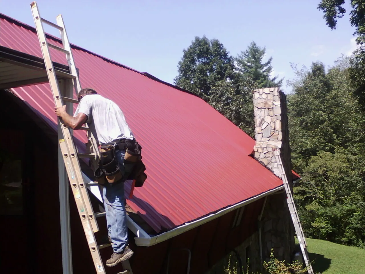 Roofer on a ladder beside a red metal roof and stone chimney — Soffit & Fascia Repair by Roofing Brothers in Hughes Springs